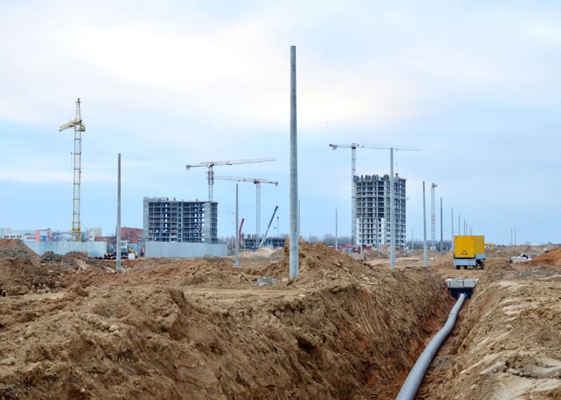 Construction site with cranes, buildings, and underground piping in progress.