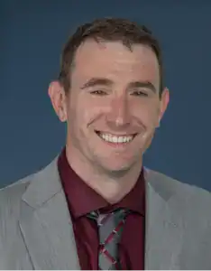 Professional portrait of Sean Kelly in a suit and tie against a blue background.